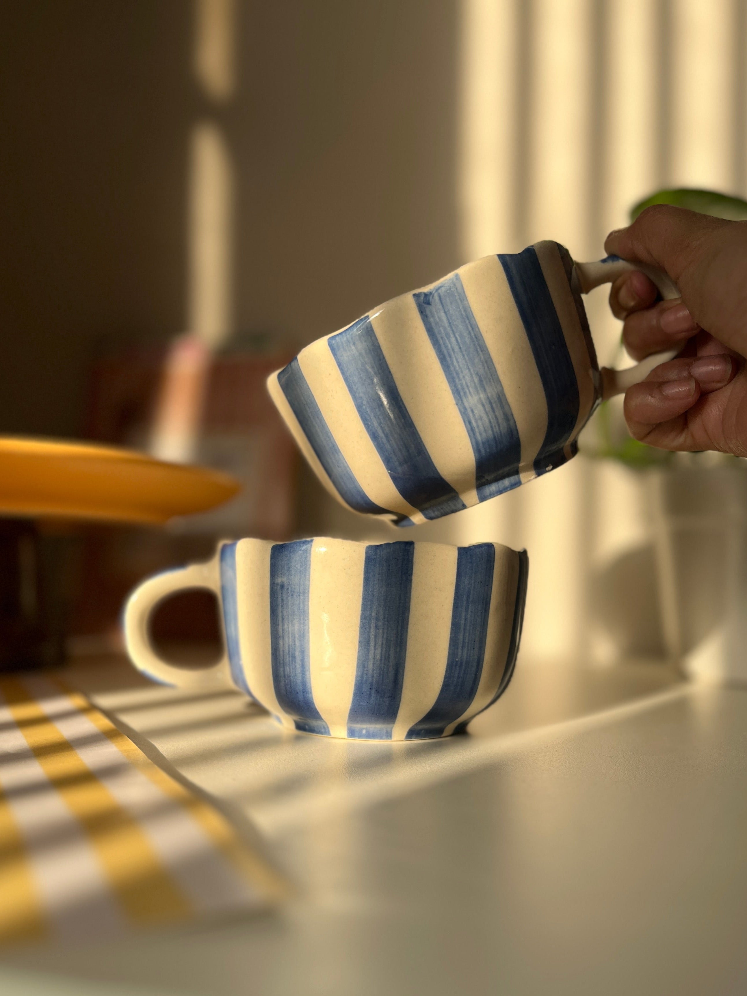 Hand holding a blue striped mug among various colorful mugs on a wooden surface. caprice striped pattern. 