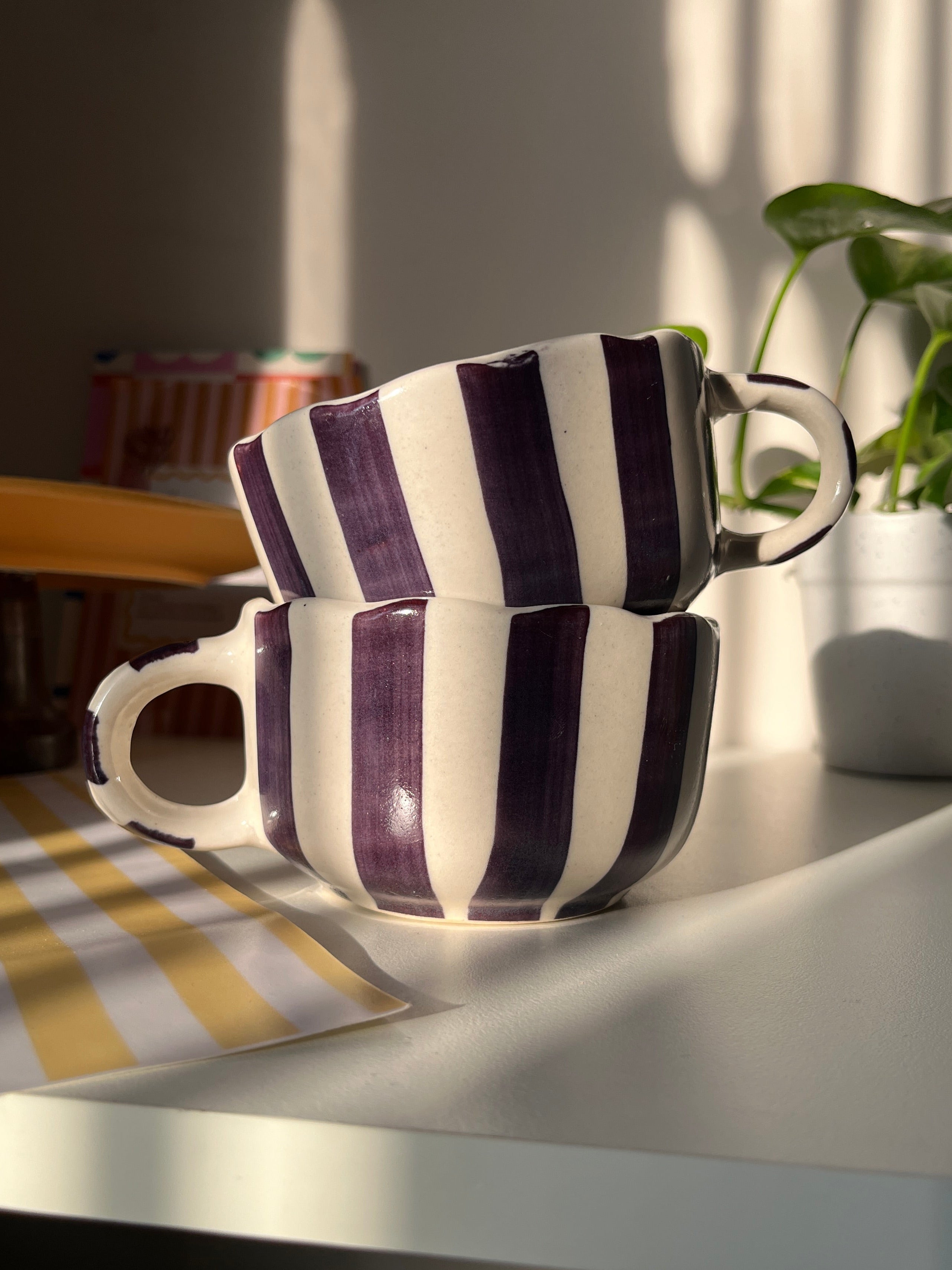 Pair of striped purple coffee and soup mugs stacked in a set of two on a hook with a plant in the background