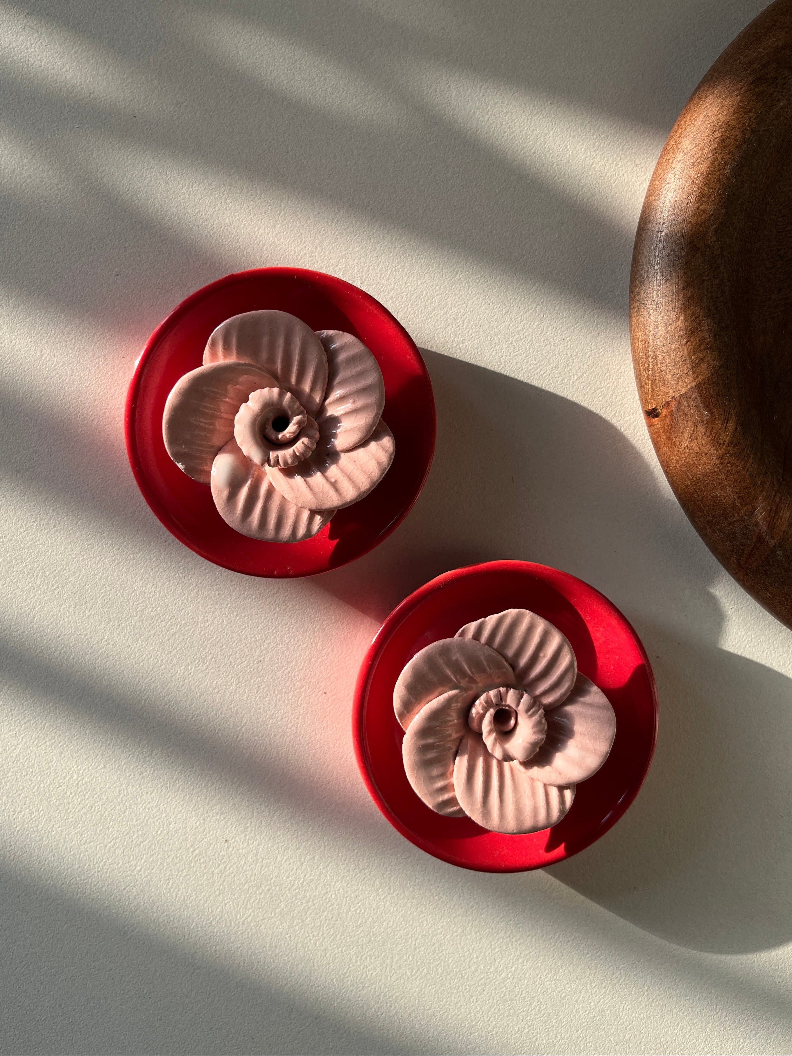 Two red ceramic flower-shaped objects on a white surface with shadows. They hold incense sticks. 