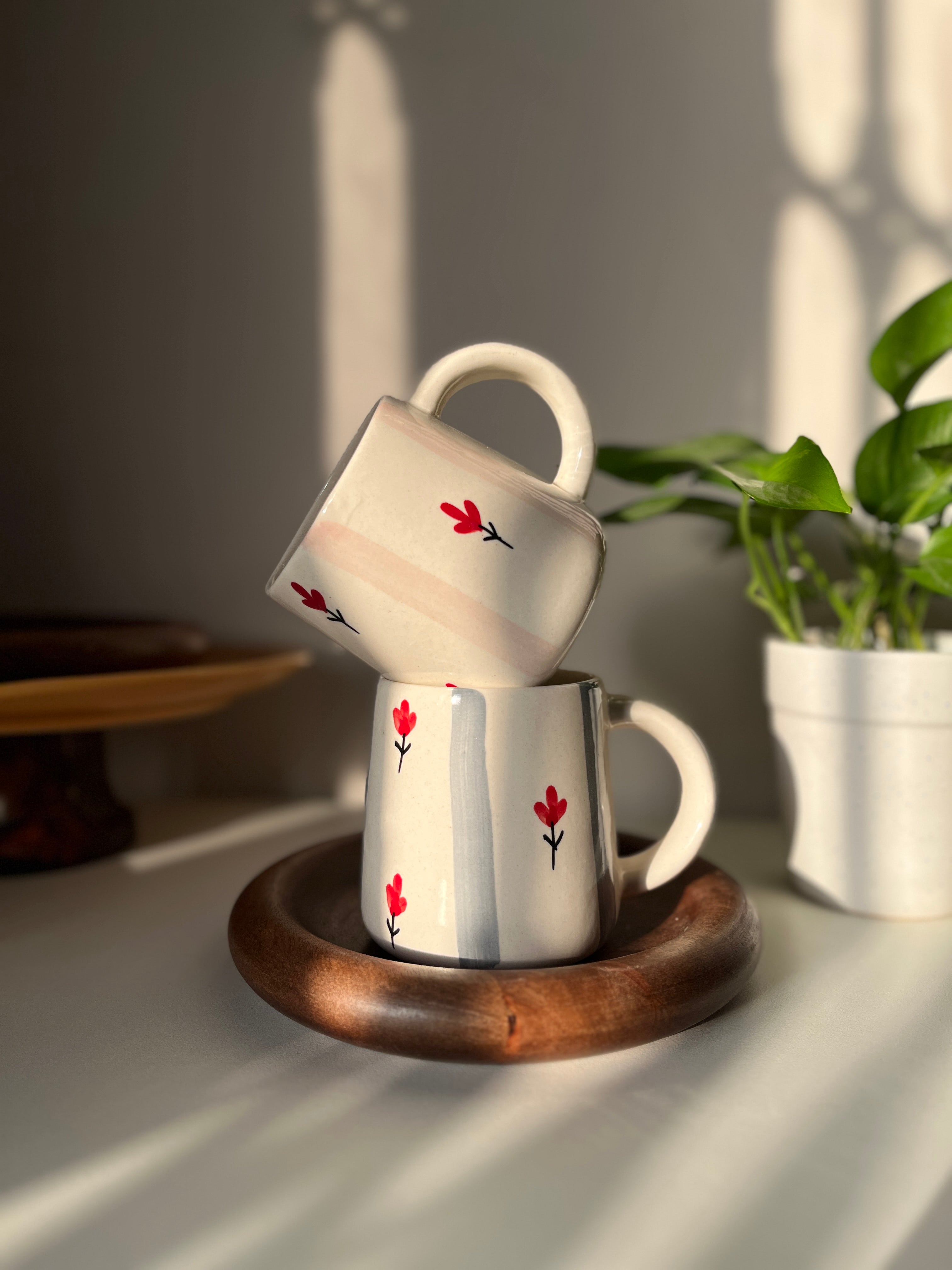 Two ceramic mugs with red flower designs on a wooden coaster, placed on a surface with a plant in the background.