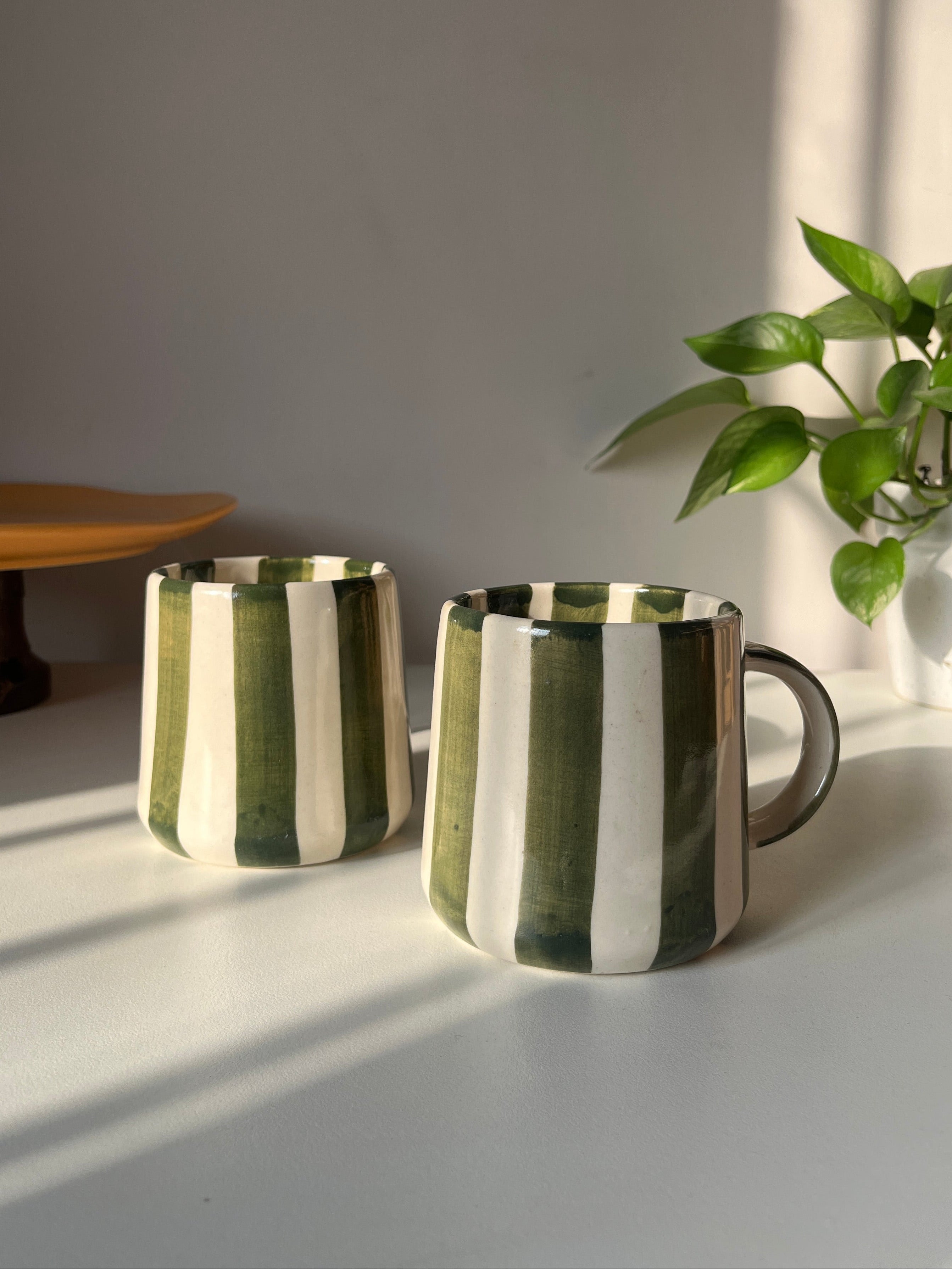 Two green striped ceramic coffee mugs on a light surface with a plant in the background