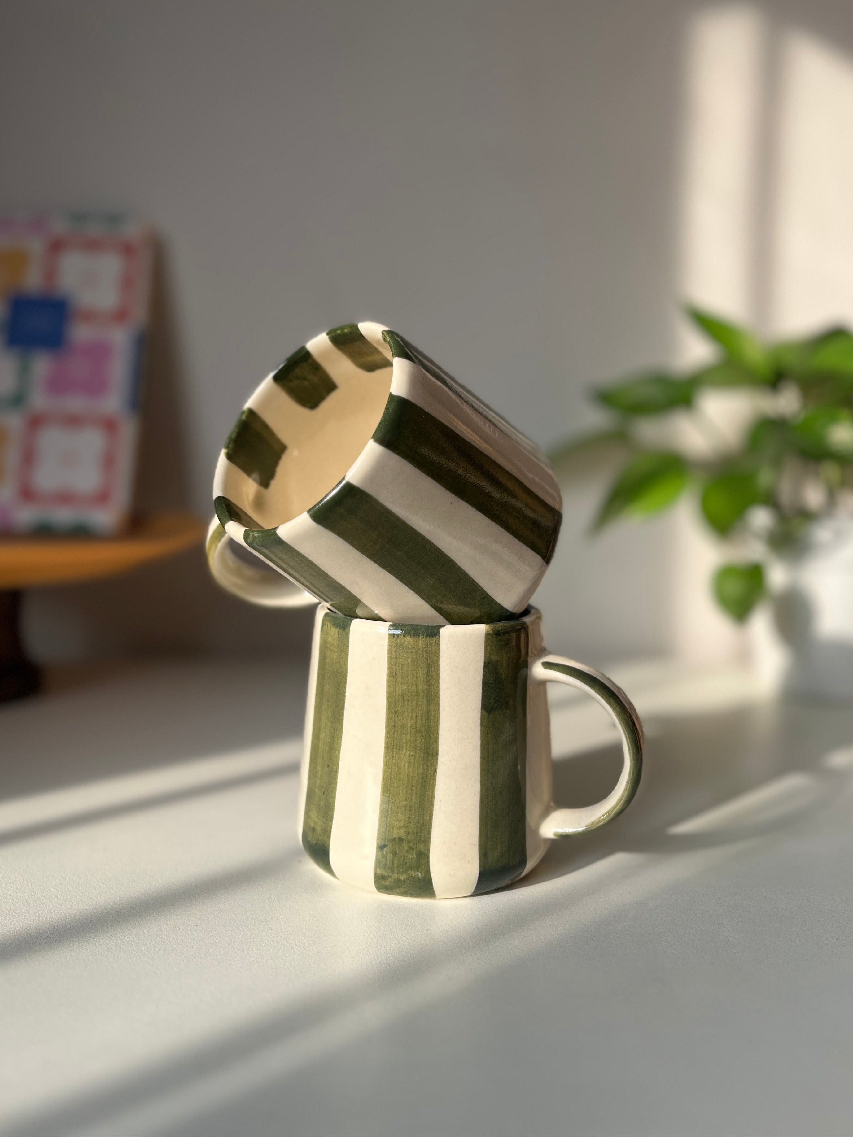 Two ceramic mugs stacked on a light surface with a plant and colorful box in the background.