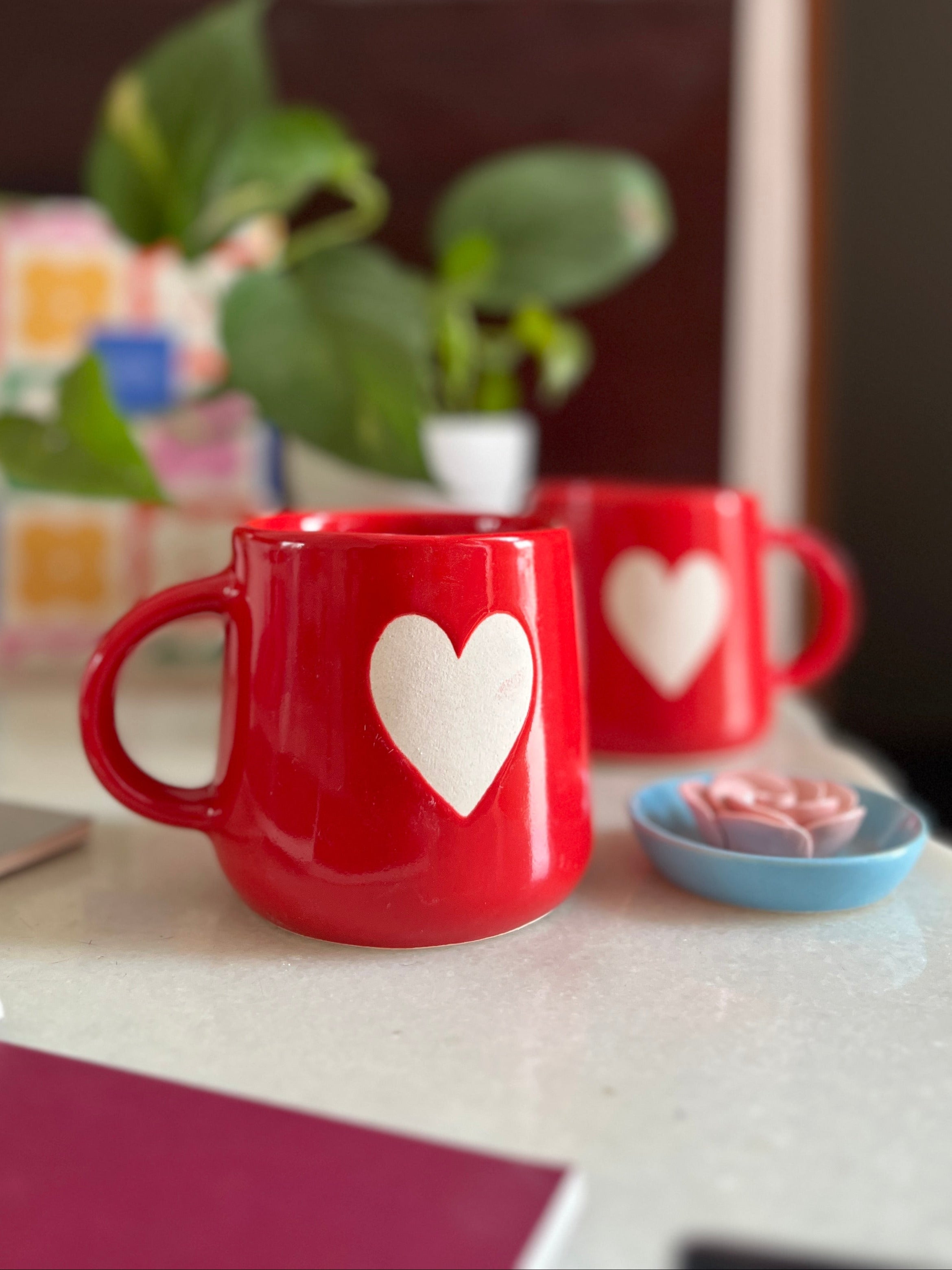 Red mugs with white hearts on a table with a plant and books in the background.