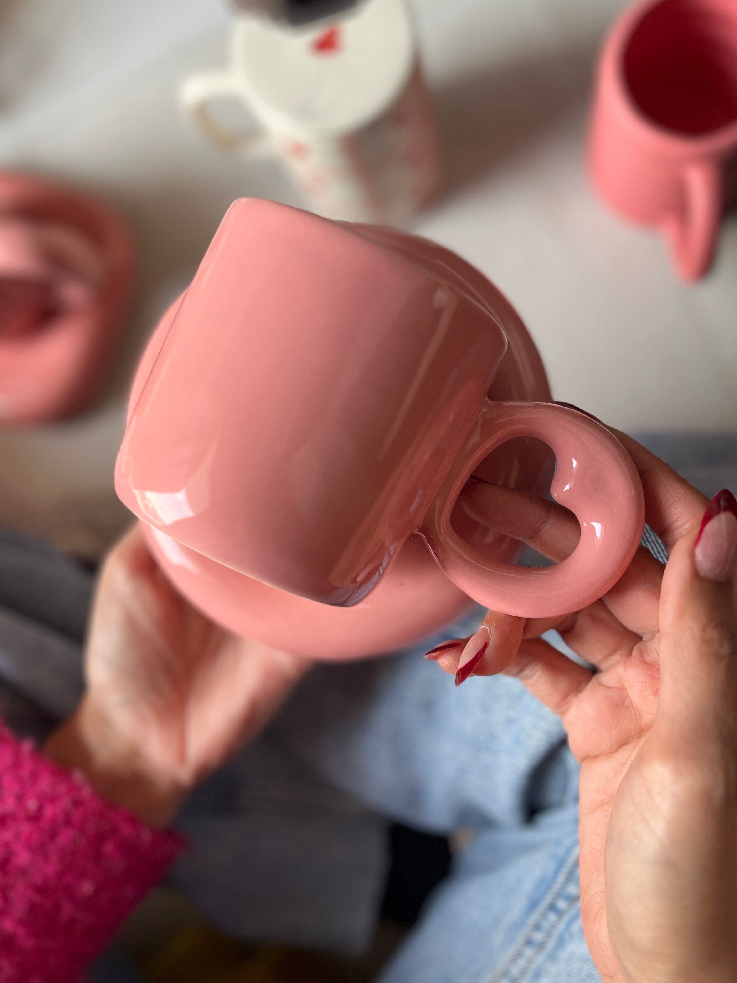 Pink ceramic cup with a round saucer. The handle of the mug is heart shaped. 