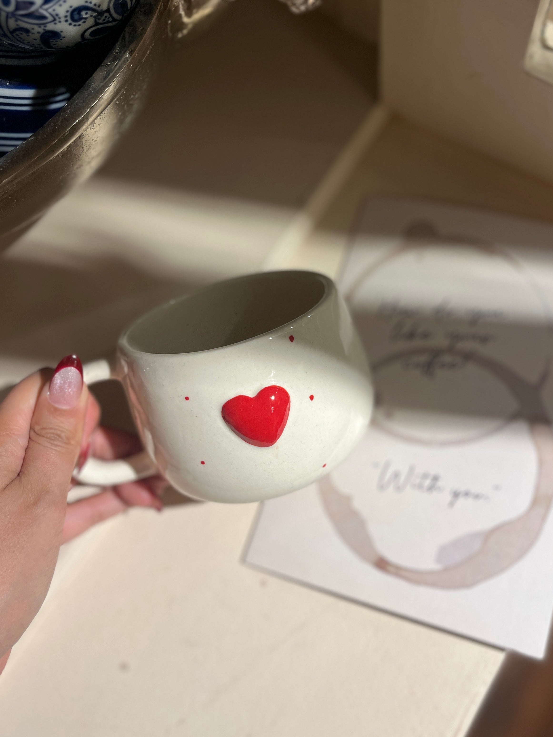 Person holding a white mug with a red heart design on a surface with a card and decorative item.