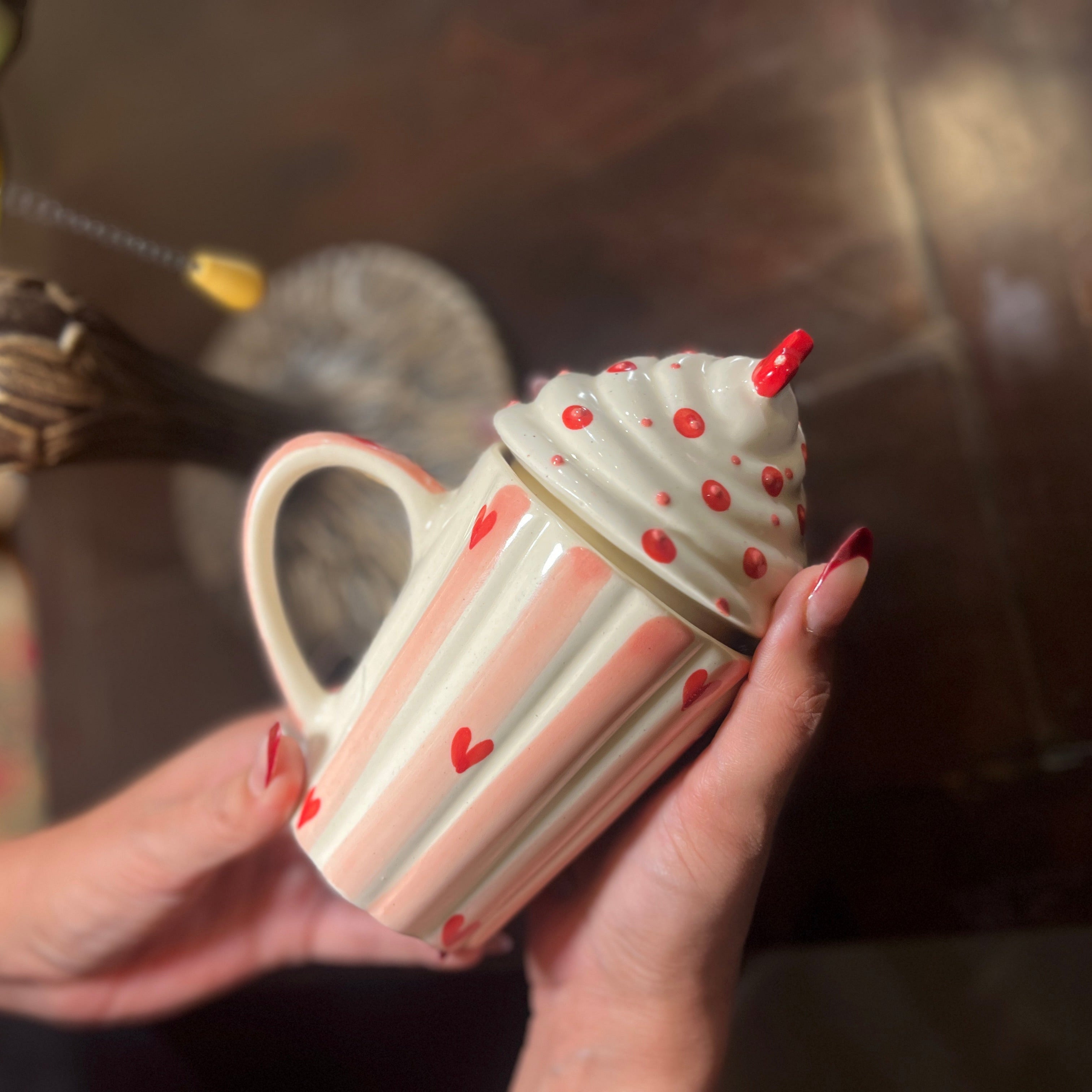 Person holding a decorative cupcake-shaped mug with red polka dots on a wooden surface.