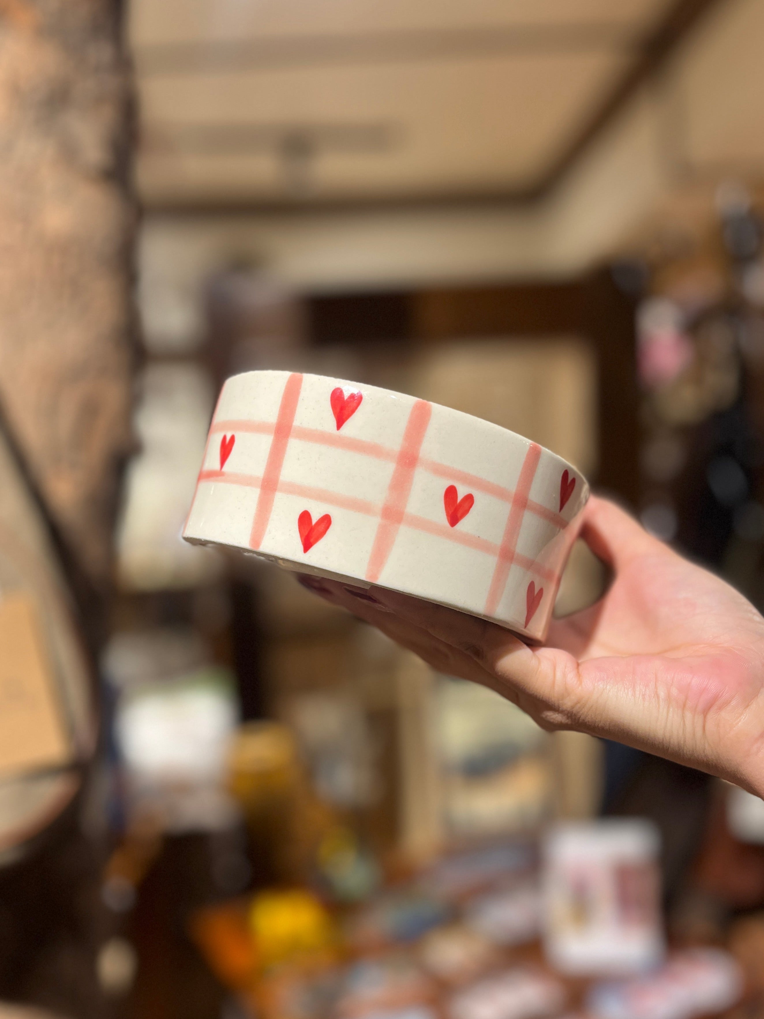 Ceramic cup with red heart pattern held by a hand in a blurred indoor setting
