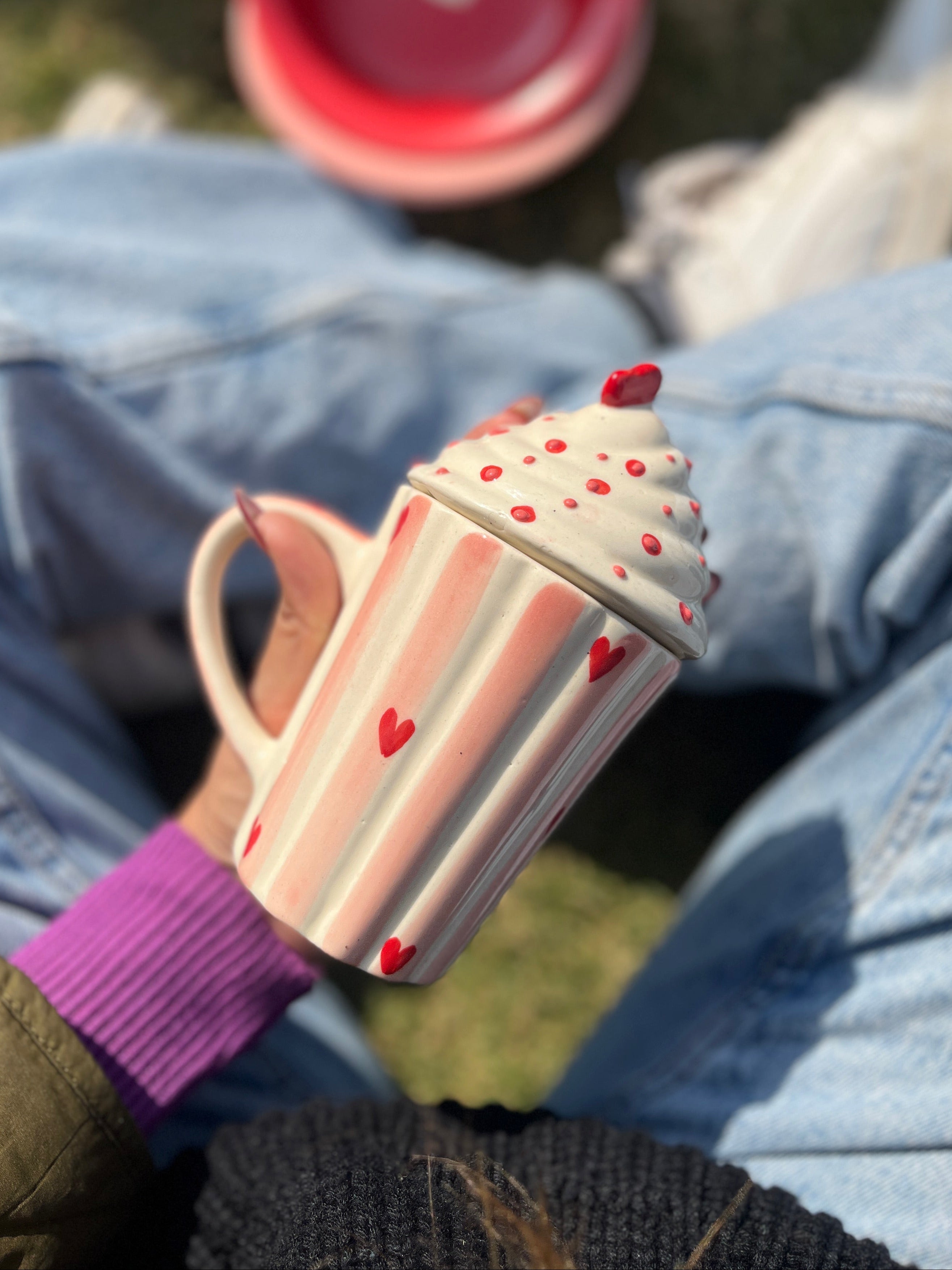 Cupcake-shaped mug with pink handle held by a person wearing a purple sleeve, sitting on blue jeans.