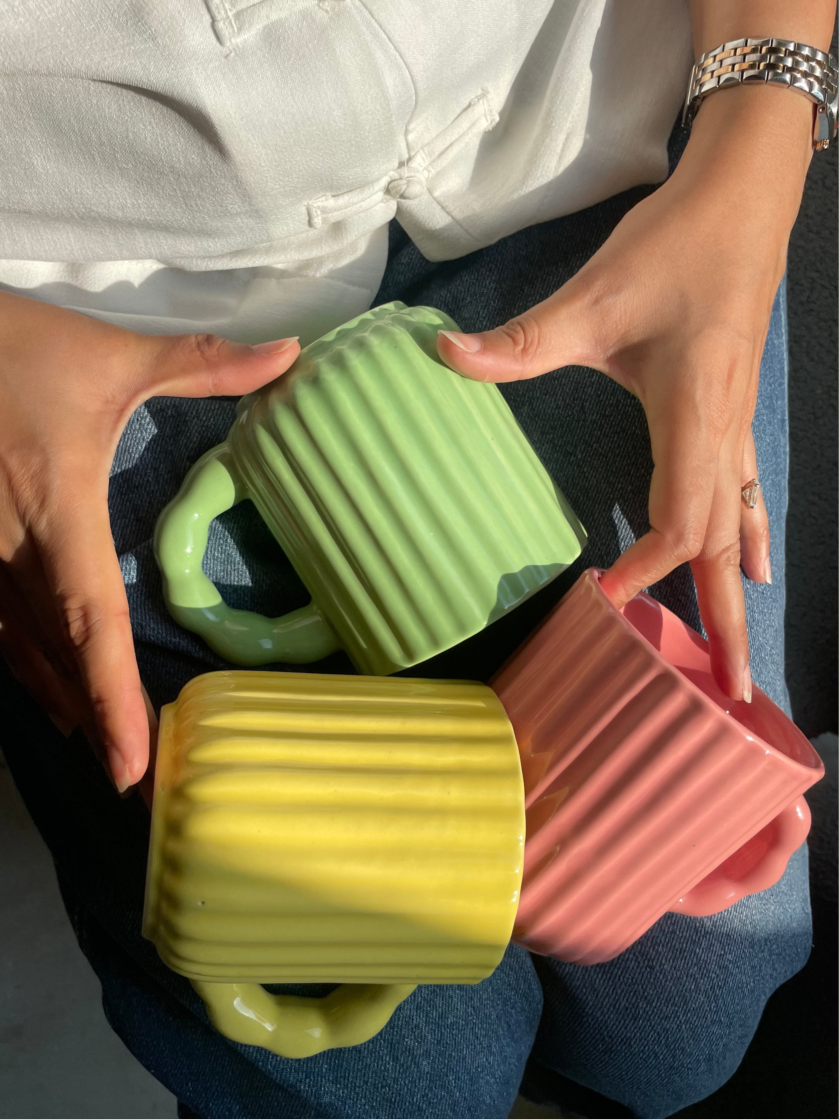 Three colorful mugs (green, yellow, pink) held by a person with a white shirt and blue jeans.