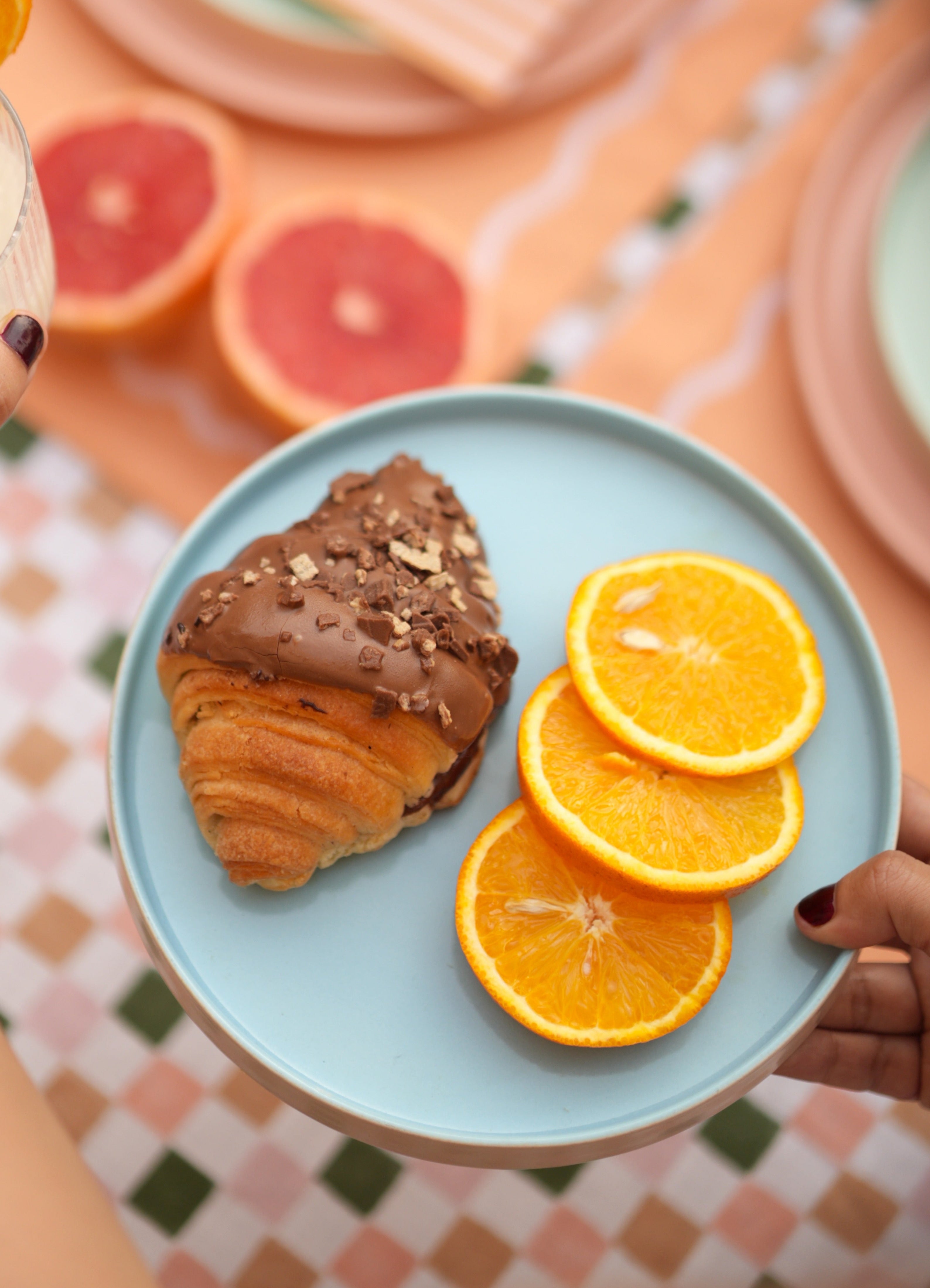 Person holding a blue ceramic pastel quarter plate with a croissant and orange slices on a colorful tablecloth.