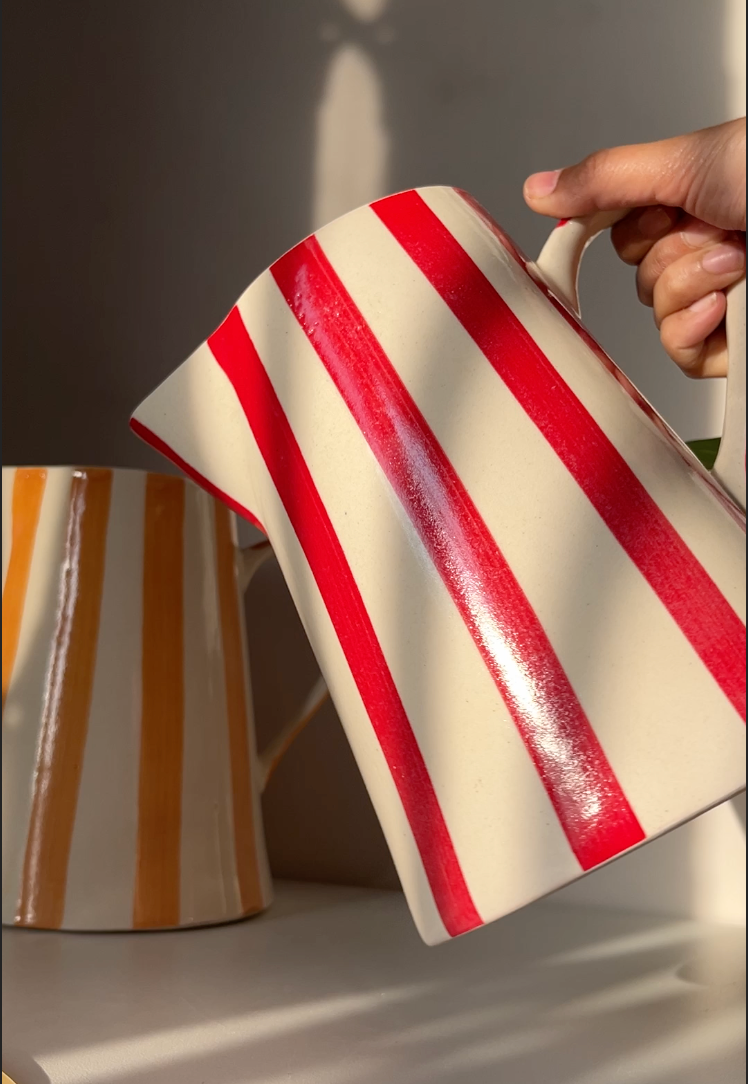 Red and white striped ceramic pitcher held by a hand with another pitcher in the background.