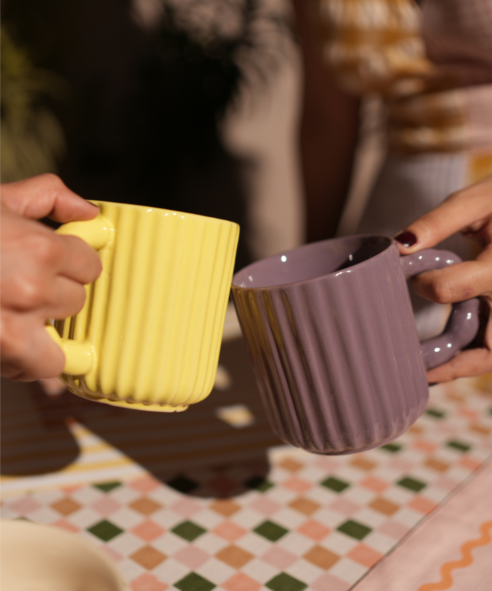 Two hands holding a yellow and a purple textured mug on a patterned tablecloth.