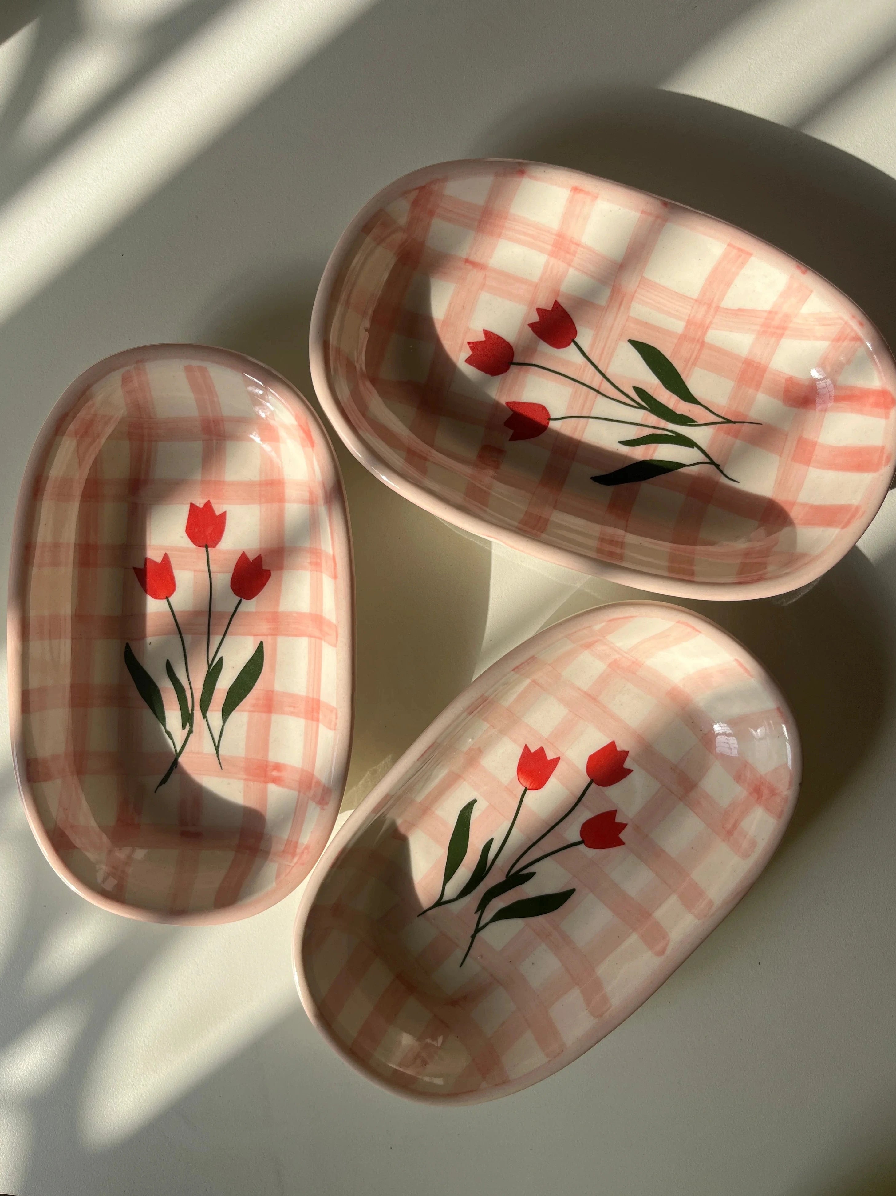 Three oval ceramic dishes with a pink and white plaid pattern and red tulip designs, arranged on a white surface with natural window shadows.
