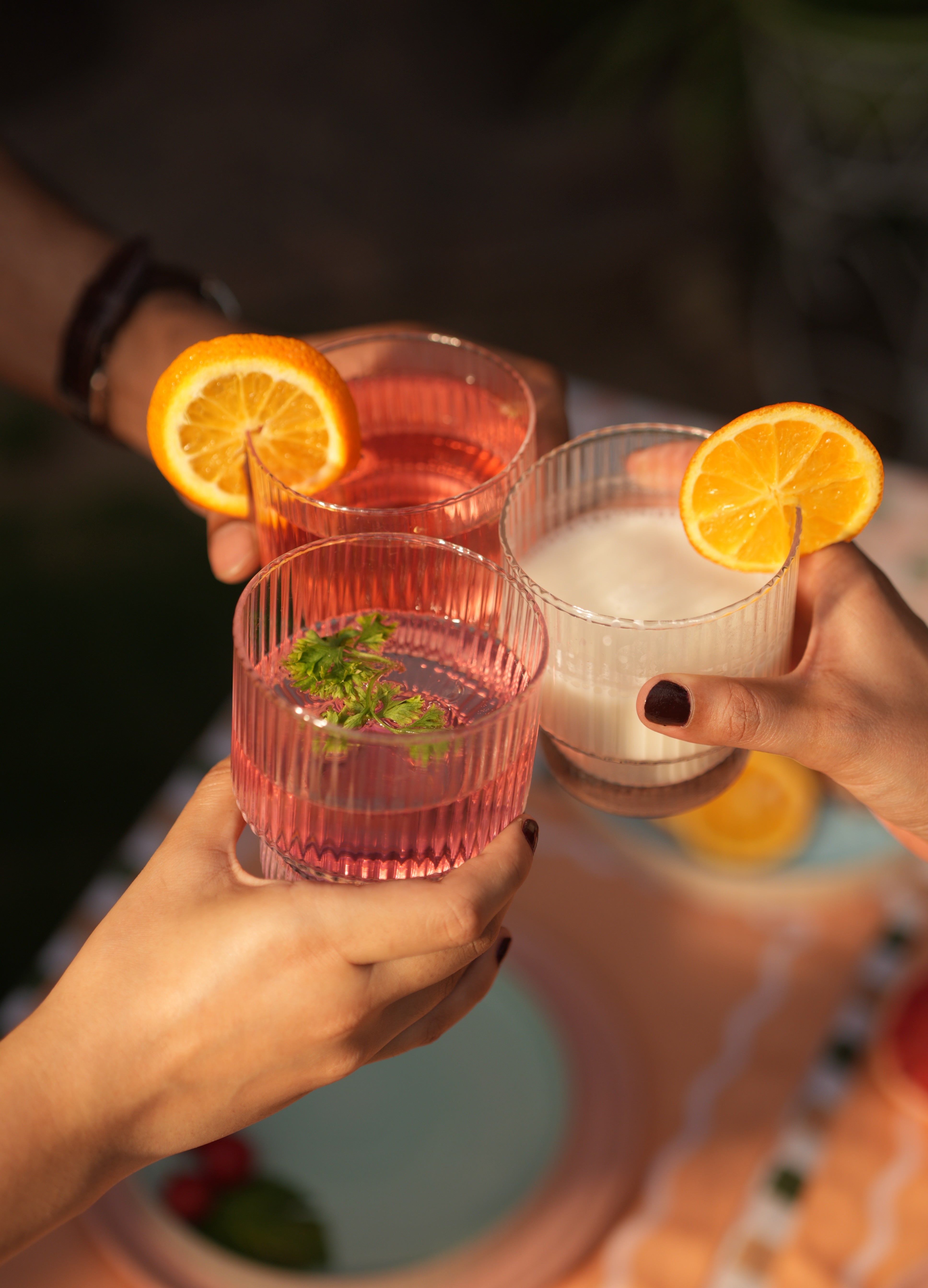 Three glasses of pink and clear drinks with orange slices held by people against a blurred background.