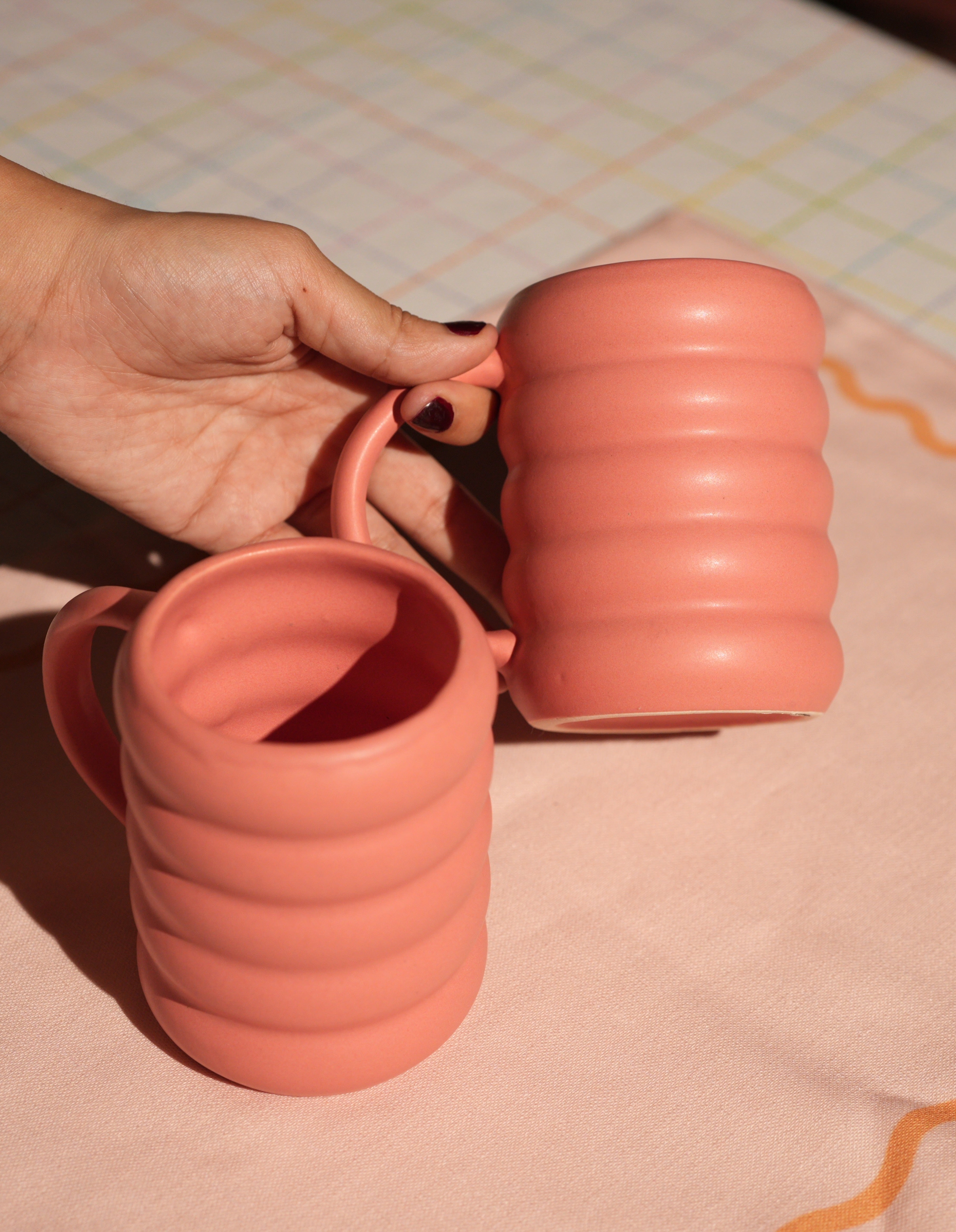 Hand holding two pink-colored ceramic tea and coffee mugs on a patterned surface