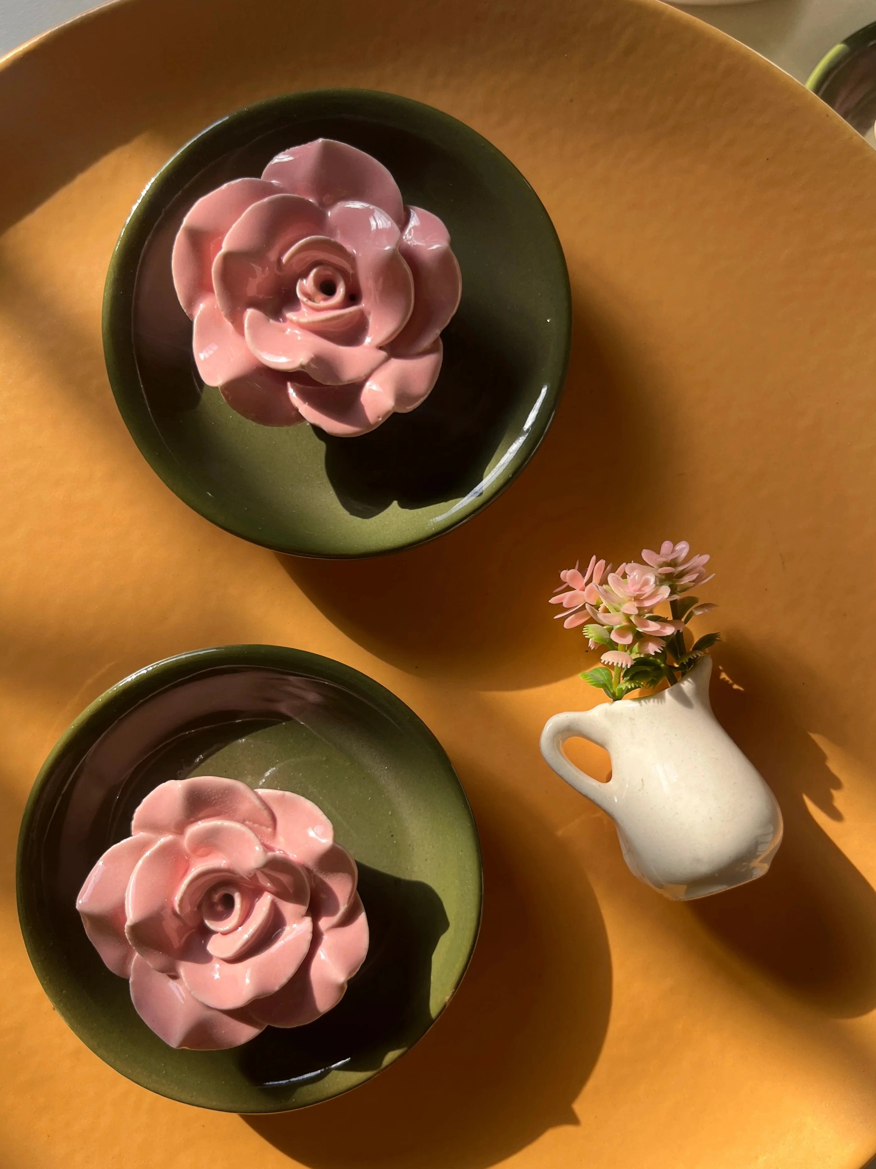 Lifestyle shot of handmade ceramic incense holder with royal green base alongside a small white vase with matching pink flowers, arranged on a large mustard-yellow tray.
