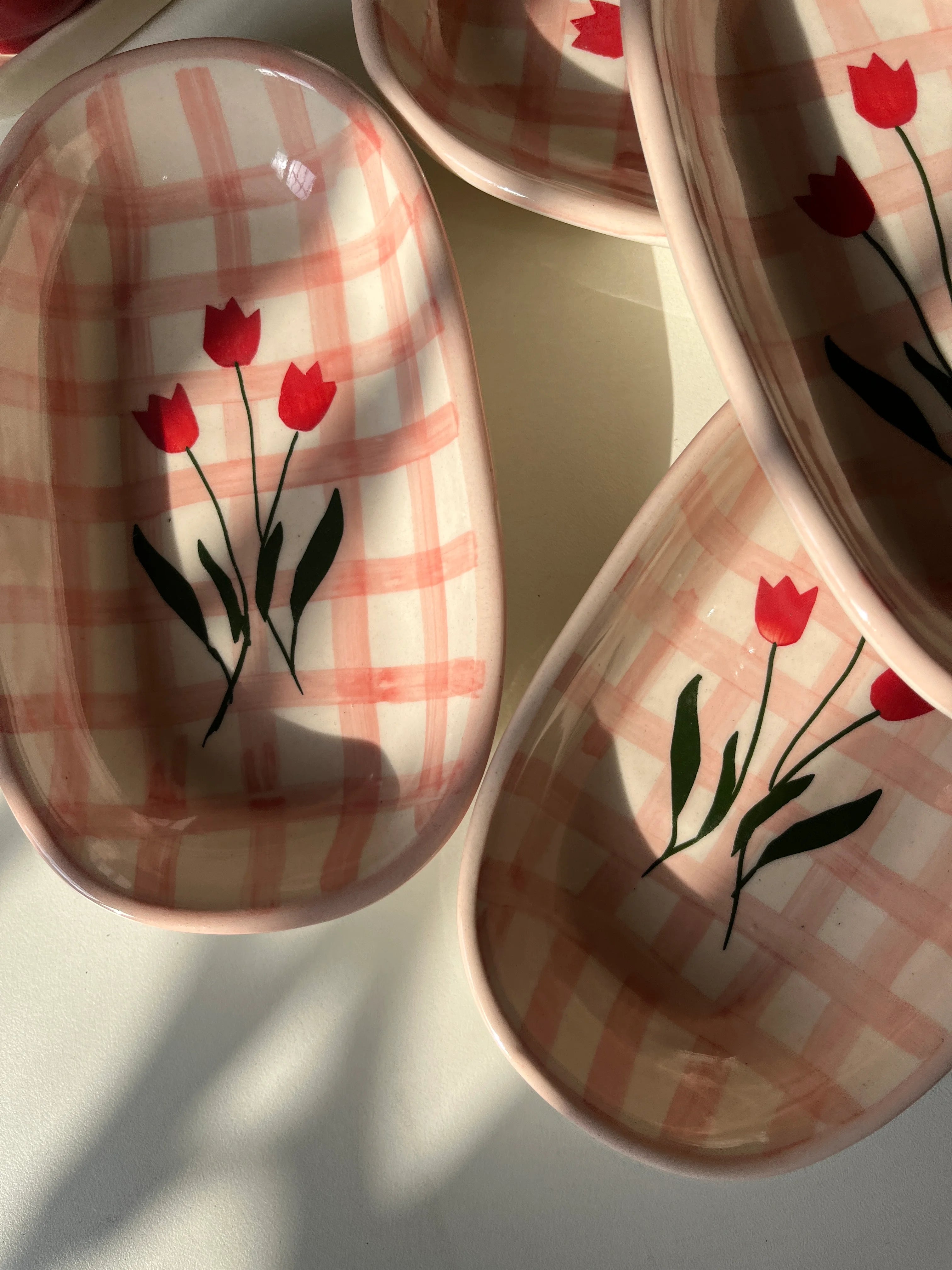 Three ceramic oval dishes with a pink and white gingham pattern and red tulip floral designs, arranged on a light surface with natural shadows.