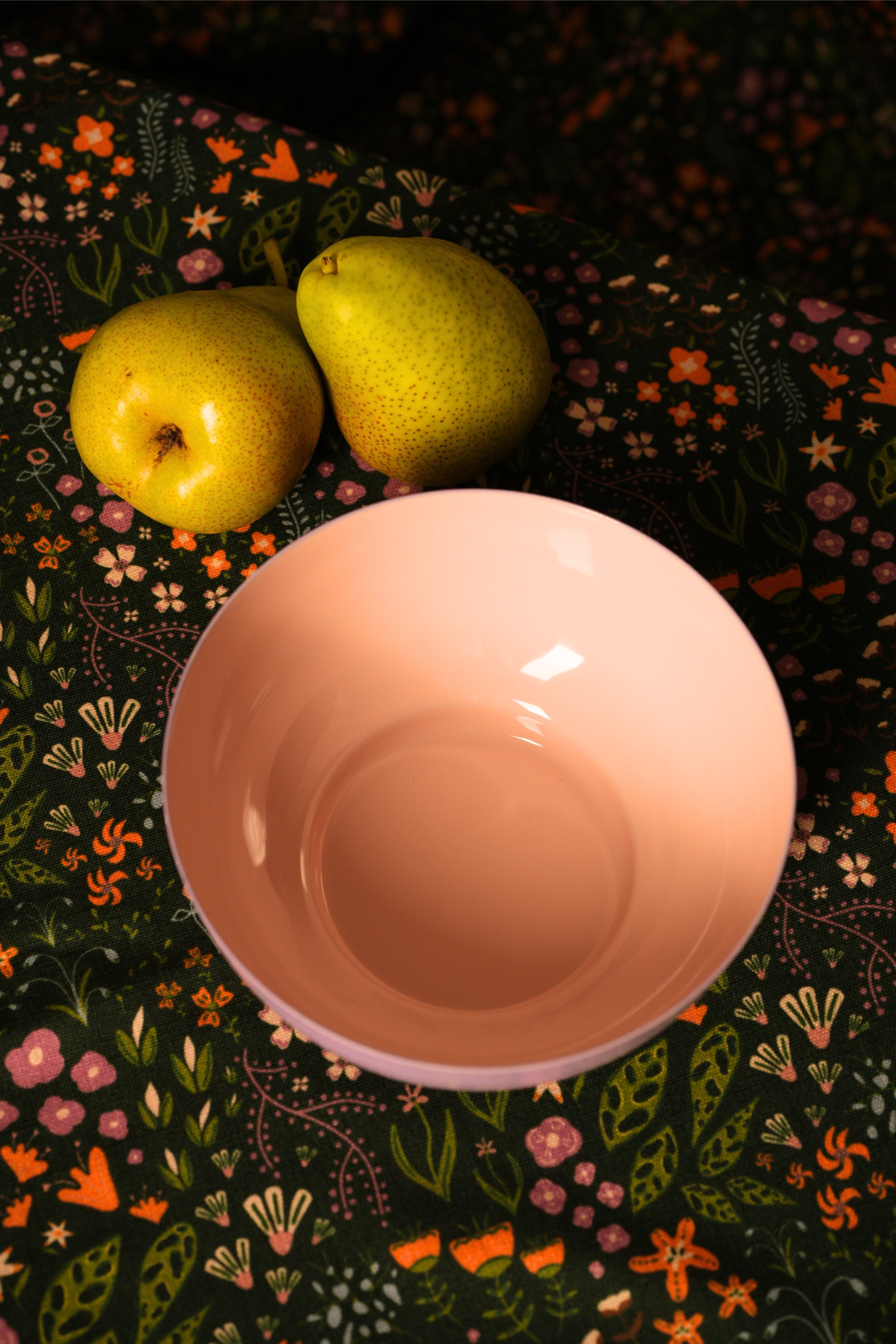 Pink metal bowl on a dark floral-patterned chihiro tablecover with two green pears.
